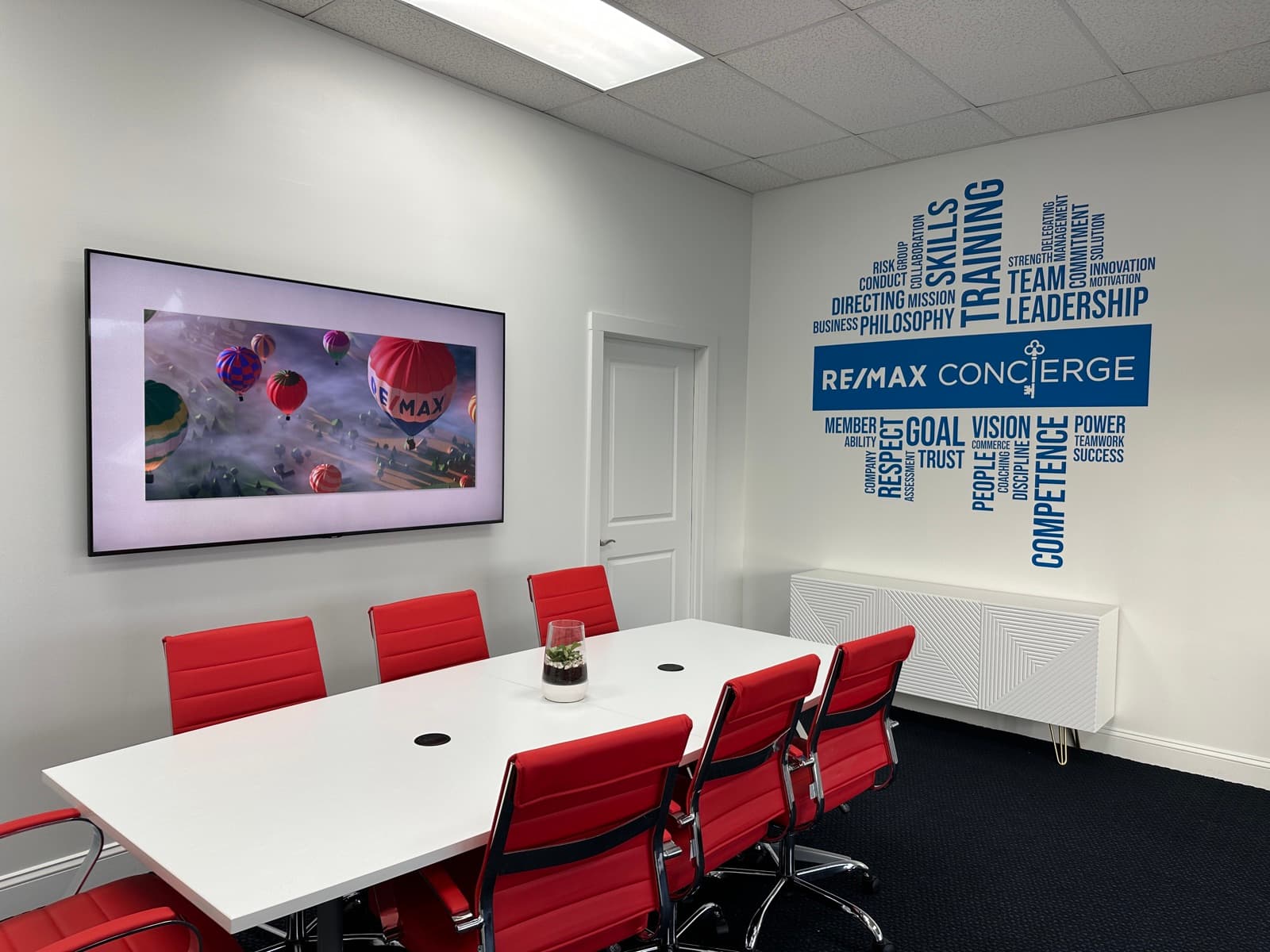 The RE/MAX Concierge conference room: a white table with red chairs, a wall-mounted screen showing RE/MAX balloons, and a wall graphic with the RE/MAX Concierge name in a blue panel, the key standing in for the i, surrounded by words including skills, training, team and leadership.